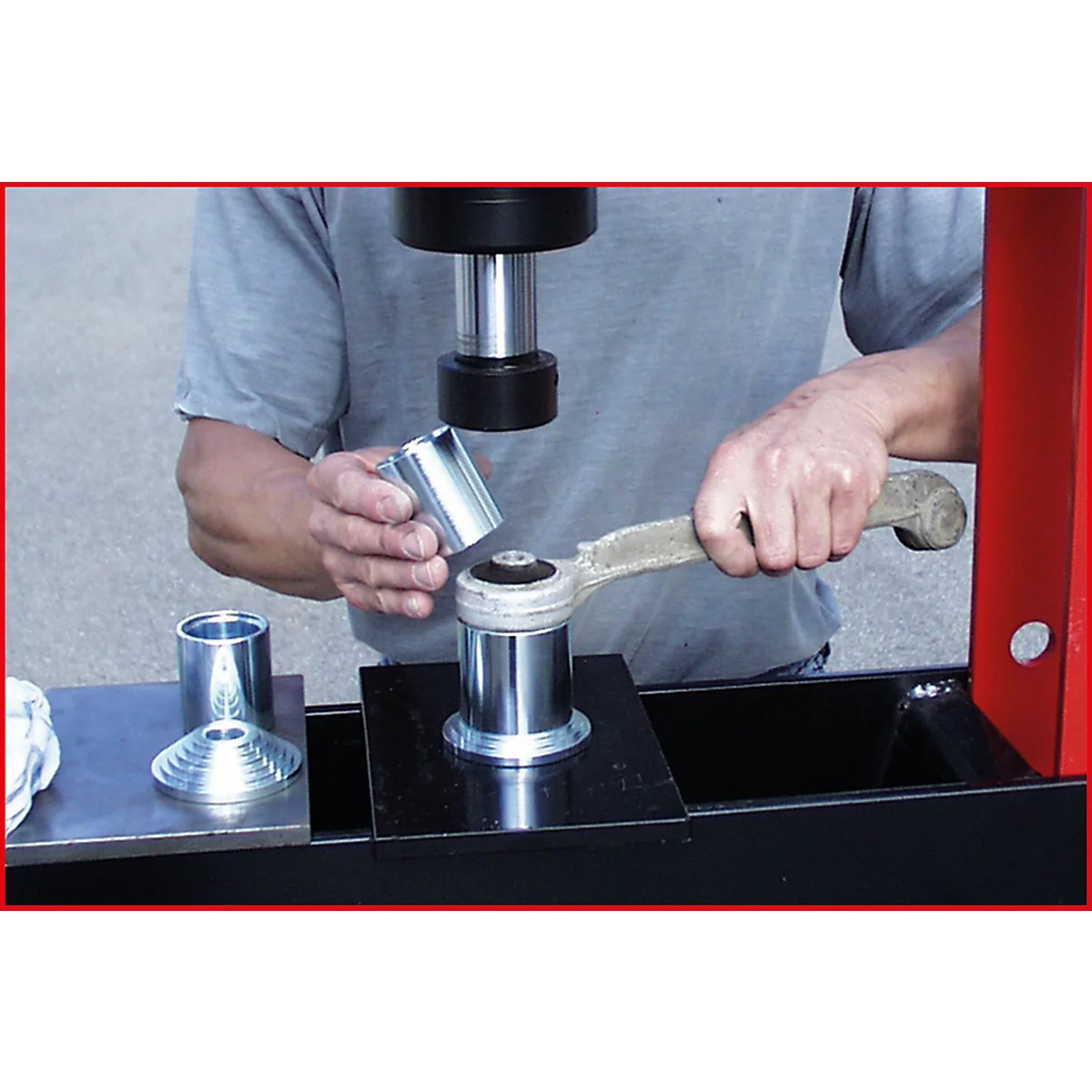 Ein Mann arbeitet mit einer Presse an einem Fahrzeugteil. Er hält einen metallischen Zylinder über ein anderes Teil. Der Hintergrund ist unscharf.